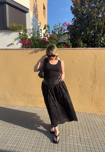 Top corto negro, falda maxi negra bordada, bolso de cintura negro con tachuelas, chanclas negras. Entorno exterior soleado con flores.