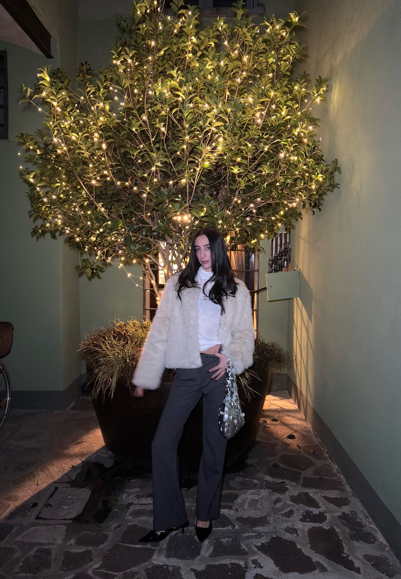 Veste en fausse fourrure blanche, haut blanc ajusté, pantalon gris évasé et sac à main pailleté. Arbre vert brillamment éclairé en arrière-plan, sol en pierre.