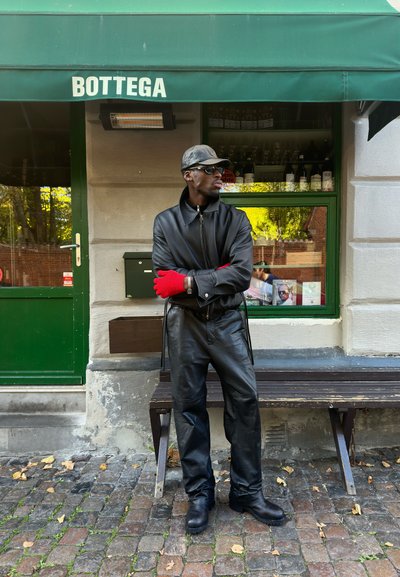Hombre con chaqueta de cuero negro, pantalones, botas, gorra y gafas de sol, de pie con los brazos cruzados y guantes rojos, frente a un café con toldo verde que dice "BOTTEGA."