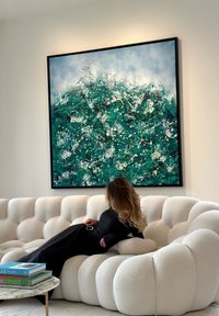 Γυναίκα με μακριά μαλλιά που ξαπλώνει σε έναν λευκό καναπέ με καπιτονέ επένδυση κάτω από έναν μεγάλο αφηρημένο πίνακα με πράσινα, λευκά και μπλε υφασμένα μοτίβα.