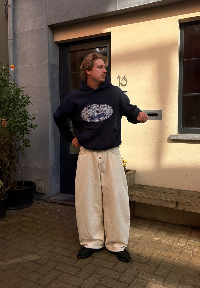 Homme en sweat à capuche noir et pantalon blanc large debout devant une maison avec une porte noire et un banc en bois, regardant sur le côté.