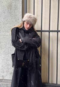 Γυναίκα με μαύρο παλτό και κασκόλ, μπεζ γούνινο καπέλο, κρατώντας μαύρη τσάντα Yves Saint Laurent, στέκεται μπροστά σε έναν τοίχο με μαύρες μεταλλικές μπάρες ενώ πέφτει χιόνι.