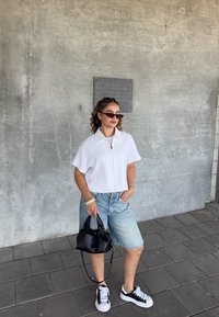 Camicia bianca a maniche corte con zip, pantaloni bermuda in denim azzurro, borsa a tracolla nera e sneakers in bianco e nero su pavimentazione grigia.