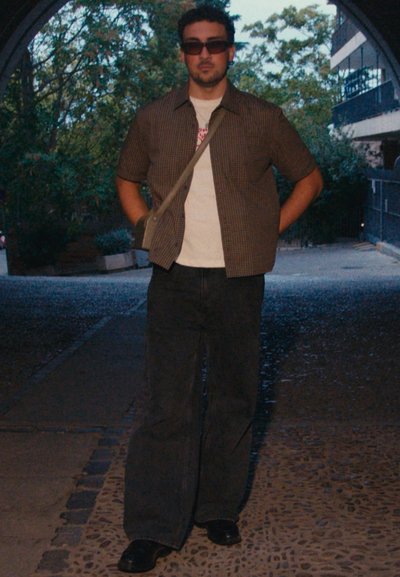 Camisa de cuadros de manga corta sobre una camiseta blanca, pantalones oscuros anchos y zapatos negros. Gafas de sol negras y un bolso bandolera completan el atuendo.