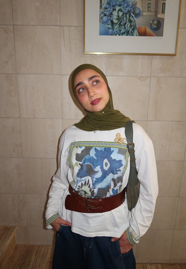 Jeune femme avec hijab vert, haut blanc à motif floral bleu, ceinture marron et jeans, debout contre un mur carrelé avec une œuvre encadrée au-dessus.