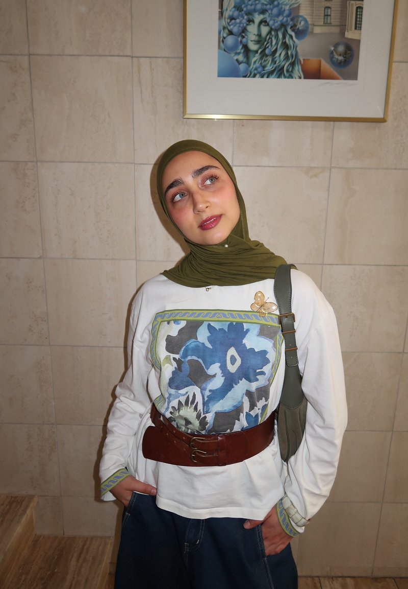 Jeune femme avec hijab vert, haut blanc à motif floral bleu, ceinture marron et jeans, debout contre un mur carrelé avec une œuvre encadrée au-dessus.