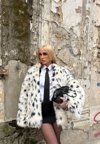 Jeune femme aux longs cheveux blonds portant un manteau en fourrure blanche à pois, une chemise blanche, une cravate noire et une jupe, tenant un sac à main noir contre un mur usé.