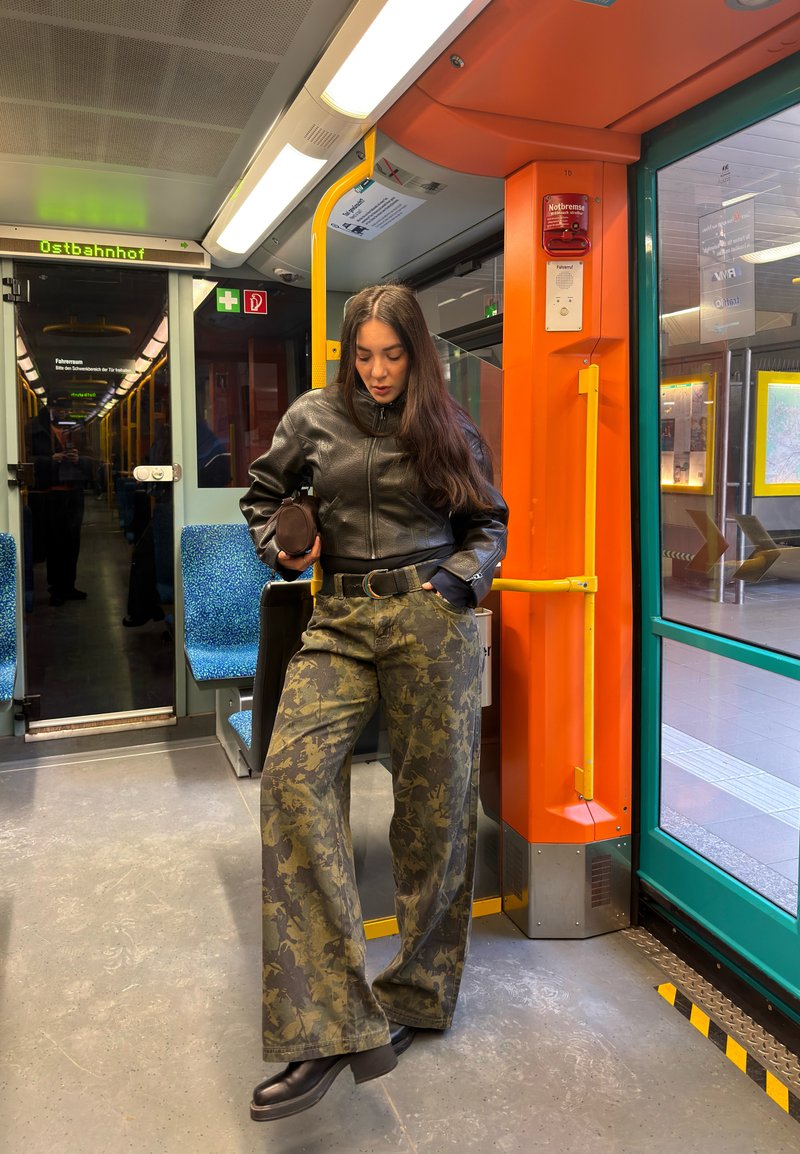 Jeune femme portant une veste en cuir et un pantalon camouflage debout à l'intérieur d'un tram près du poteau orange et des sièges bleus à la station Ostbahnhof.