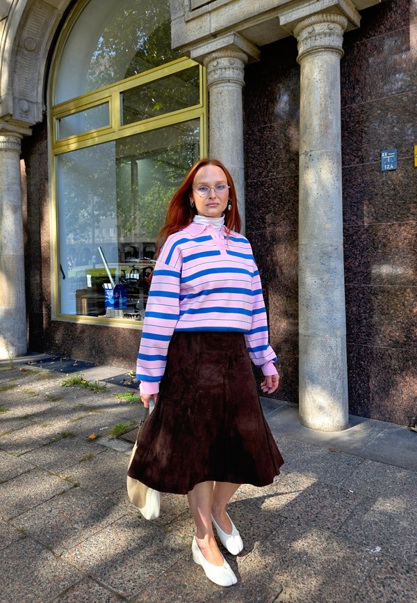 Une femme rousse portant des lunettes pose devant un bâtiment aux colonnes de pierre. Elle porte un haut rayé rose et bleu, une jupe foncée et des chaussures blanches.