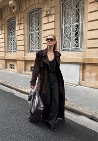 Μακριά καφέ σουέντ παλτό, μαύρη μπλούζα, φαρδιά παντελόνια και μυτερές μαύρες γόβες. Κρατάει μια μεγάλη γκρι δερμάτινη τσάντα. Σε αστική τοποθεσία.