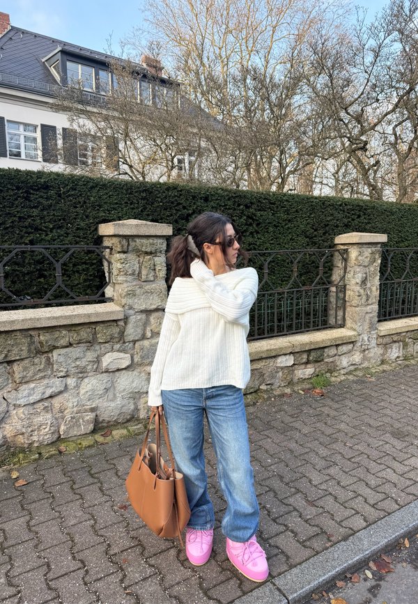 White ribbed jumper, light blue baggy jeans, and pink shoes. Brown tote bag. Urban setting with stone wall and greenery in the background.