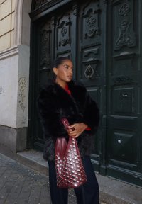 Palton din blană sintetică neagră, cu un strat interior roșu, geantă mare bordeaux cu ținte argintii, blugi închisi la culoare, stând în fața unei uși verzi. Texturi netede.