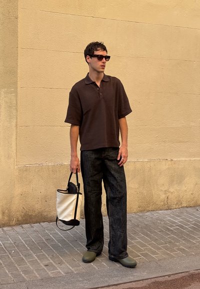 Camisa polo marrón de manga corta, pantalones de mezclilla oscuros, zapatos verdes tipo slip-on, y una bolsa tote ligera bicolor. De pie contra una pared beige.