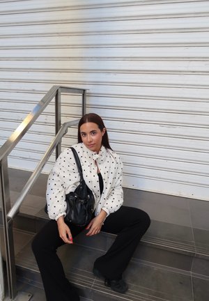 Mujer con chaqueta blanca de lunares y pantalones negros sentada en las escaleras junto a una barandilla metálica, sosteniendo un bolso negro.