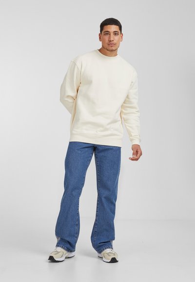 Sudadera de cuello redondo color crema con un ajuste relajado, combinada con jeans rectos azules y zapatillas deportivas de color claro. Diseño sencillo, fabricada en una mezcla de algodón.