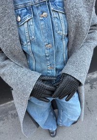 Personne portant un manteau en laine gris par-dessus une veste en denim boutonnée, un jean bleu, des gants en cuir tressé noirs et des chaussures noires, assise à l'extérieur sur le trottoir.