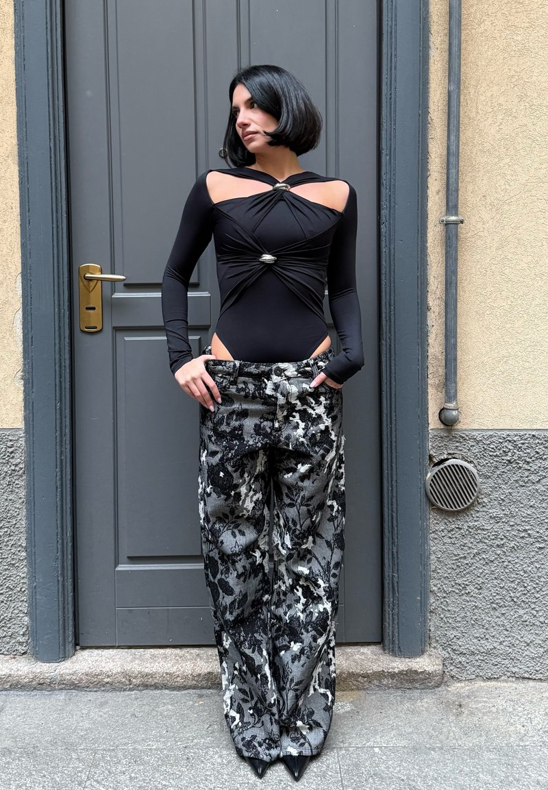 Body de manga comprida preto com recortes e ferragens prateadas, combinado com calças largas pretas com padrão floral branco e sapatos pretos pontiagudos.