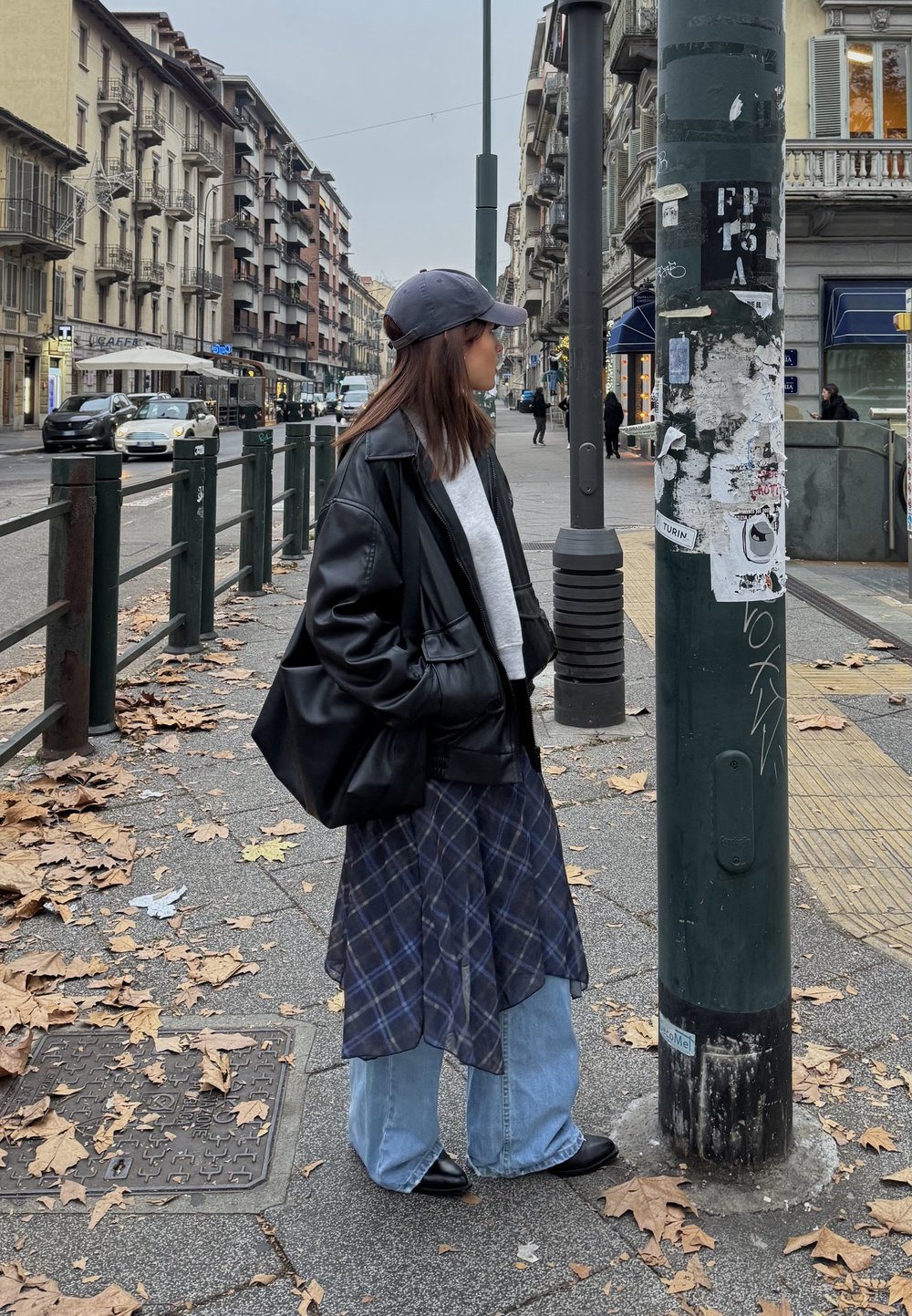 Black leather jacket, grey jumper, long checked skirt, wide denim jeans, black shoes, standing next to a green pole with stickers.
