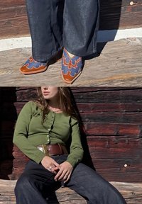 Brun- og blåbroderte sko med spisse tær; grønn cardigan med knapper; mørke vide bukser; trebakgrunn.