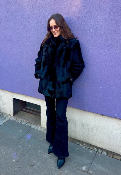 Abrigo de piel sintética negra con un cuello alto, bolsillos laterales y un acabado texturizado, llevado sobre pantalones ajustados de campana negros y tacones puntiagudos negros.