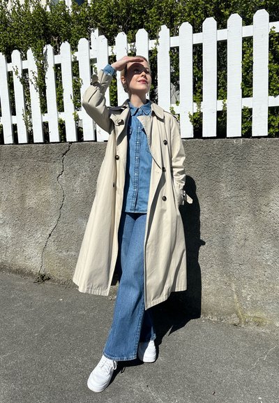 Gabardina beige sobre una camisa de mezclilla, combinada con pantalones vaqueros de pierna ancha azules y zapatillas blancas. De pie contra una pared de piedra con una cerca blanca de estacas.
