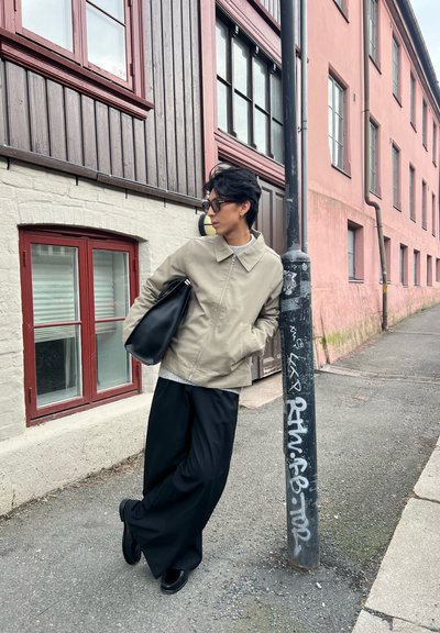 Chaqueta beige clara, suéter gris, pantalones de pierna ancha negros y zapatos negros. De pie junto a un poste cerca de un edificio con ventanas rojas.