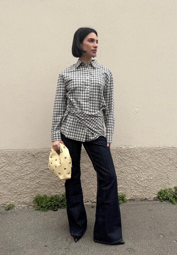 Donna con capelli neri corti, indossa una camicia a quadri e jeans scuri svasati, tiene in mano una borsa beige con borchie metalliche, in piedi davanti a una parete beige.