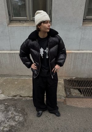 Persona que lleva un gorro blanco, chaqueta acolchada negra, camiseta negra con estampado, pantalones negros holgados y zapatos negros, de pie en una calle cerca de un edificio.