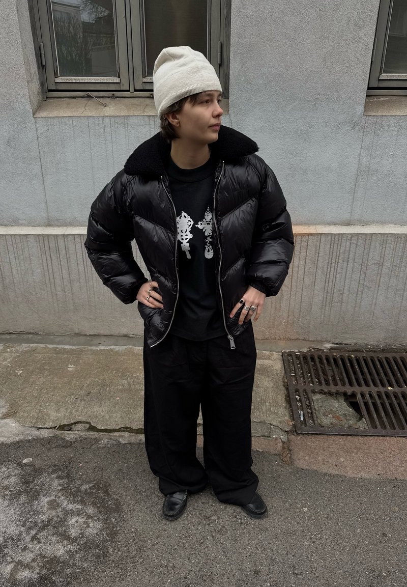 Personne portant un bonnet blanc, une veste matelassée noire, un t-shirt graphique noir, un pantalon noir ample et des chaussures noires, debout dans une rue près d'un bâtiment.
