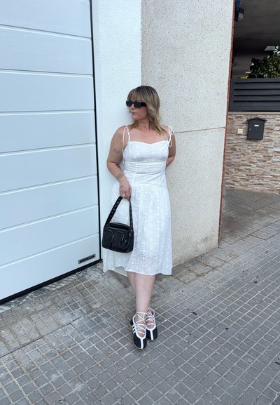 Vestido blanco de ojal con tirantes ajustables, falda fluida, combinado con un bolso negro y zapatos de cordones en blanco y negro, contra una pared texturizada.