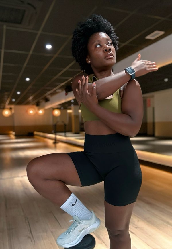 Frau in Sportkleidung streckt ihren Arm in einem Fitnessstudio mit Holzboden und Deckenbeleuchtung und balanciert auf einem kleinen schwarzen Gegenstand mit einem Fuß.