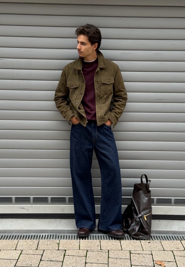 Jeune homme en veste olive, pull bordeaux, jean large foncé et chaussures marron, debout les mains dans les poches, à côté d'un sac à dos en cuir noir.