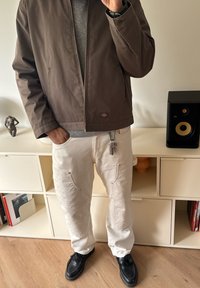 Ruskea takki harmaan neuleen päällä, yhdistettynä valkoisiin housuihin, joissa on kohopainetut tekstit taskuissa. Mustat kengät, seisoo puisella lattialla.