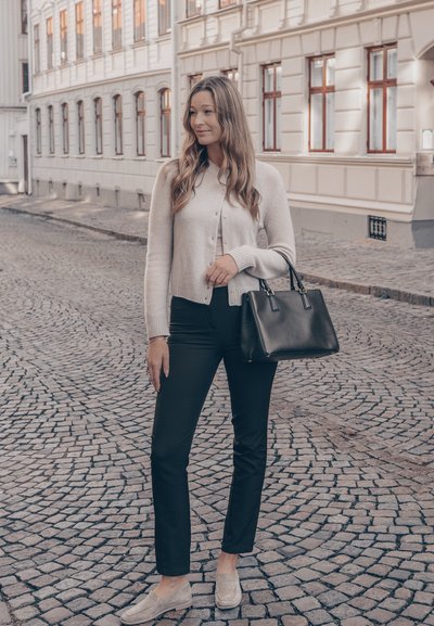 Cárdigan gris claro, pantalones negros y zapatos beige sin cordones. Bolso negro con doble asa. Calle adoquinada y edificios históricos al fondo.