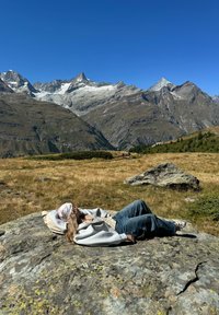 Person som ligger tilbakelent på en stein, iført en grå hettegenser, rutete skjorte og blå jeans. Omgivende fjell er delvis dekket med snø under en klar himmel.
