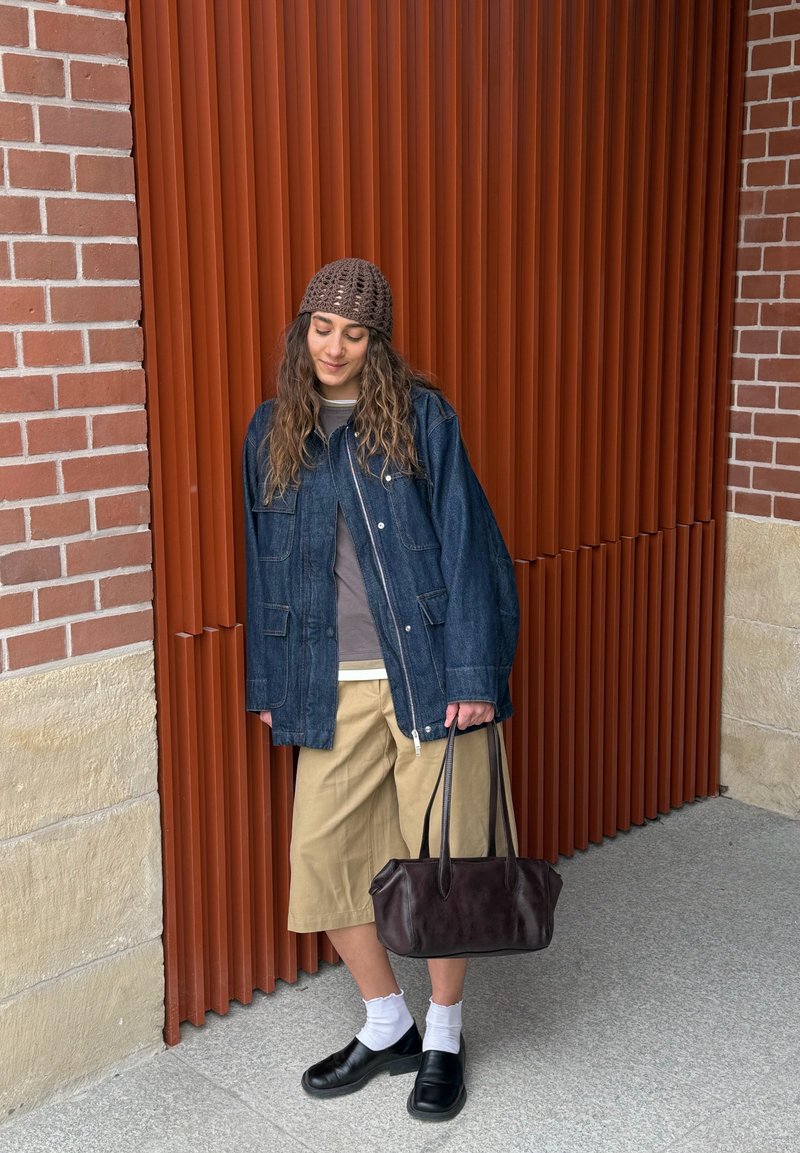 Junge Frau mit brauner Strickmütze, Jeansjacke, beigen Culottes, weißen Socken, schwarzen Schuhen, hält eine dunkelbraune Handtasche und steht vor einer roten Lattenwand.