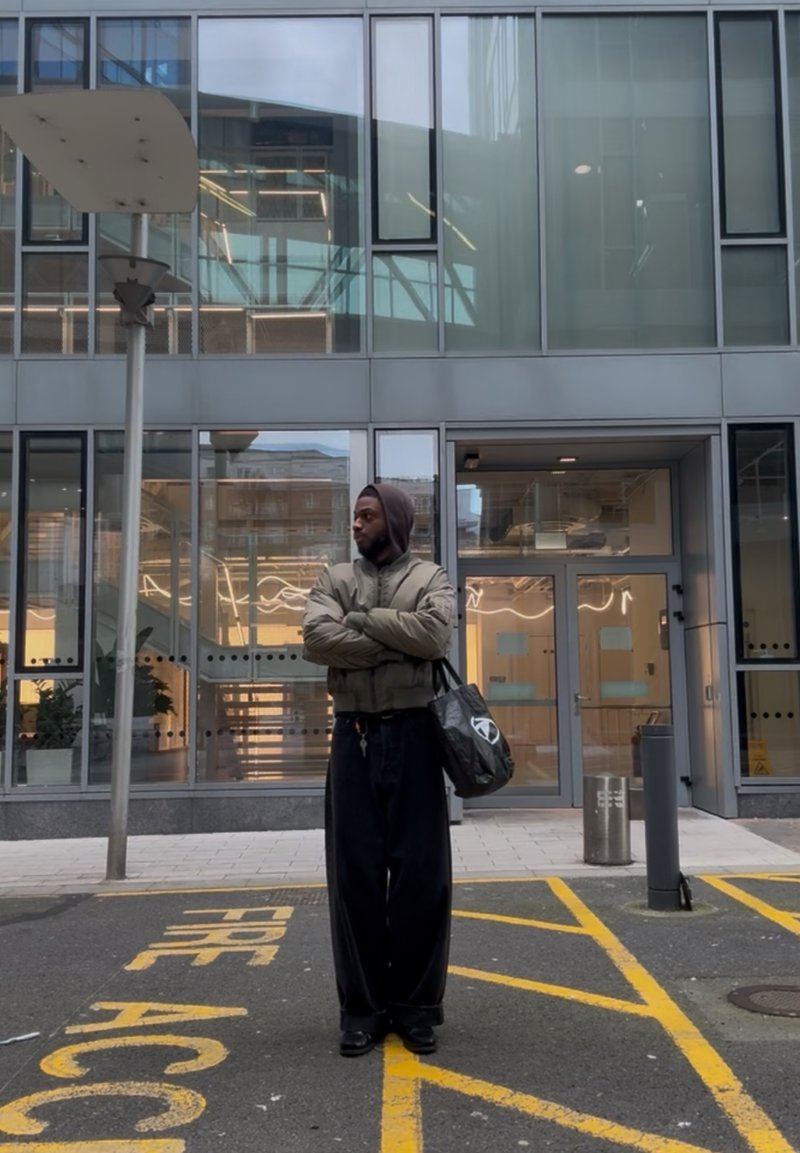 Homme portant une veste à capuche et un sac, debout les bras croisés sur une voie d'accès incendie jaune devant l'entrée d'un bâtiment en verre.