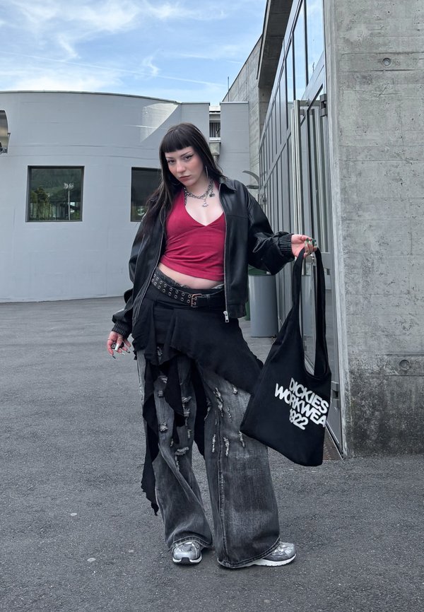 Jeune personne en jean large déchiré, crop top rouge, veste en cuir noire, baskets argentées, tenant un sac tote noir avec un texte blanc en extérieur.