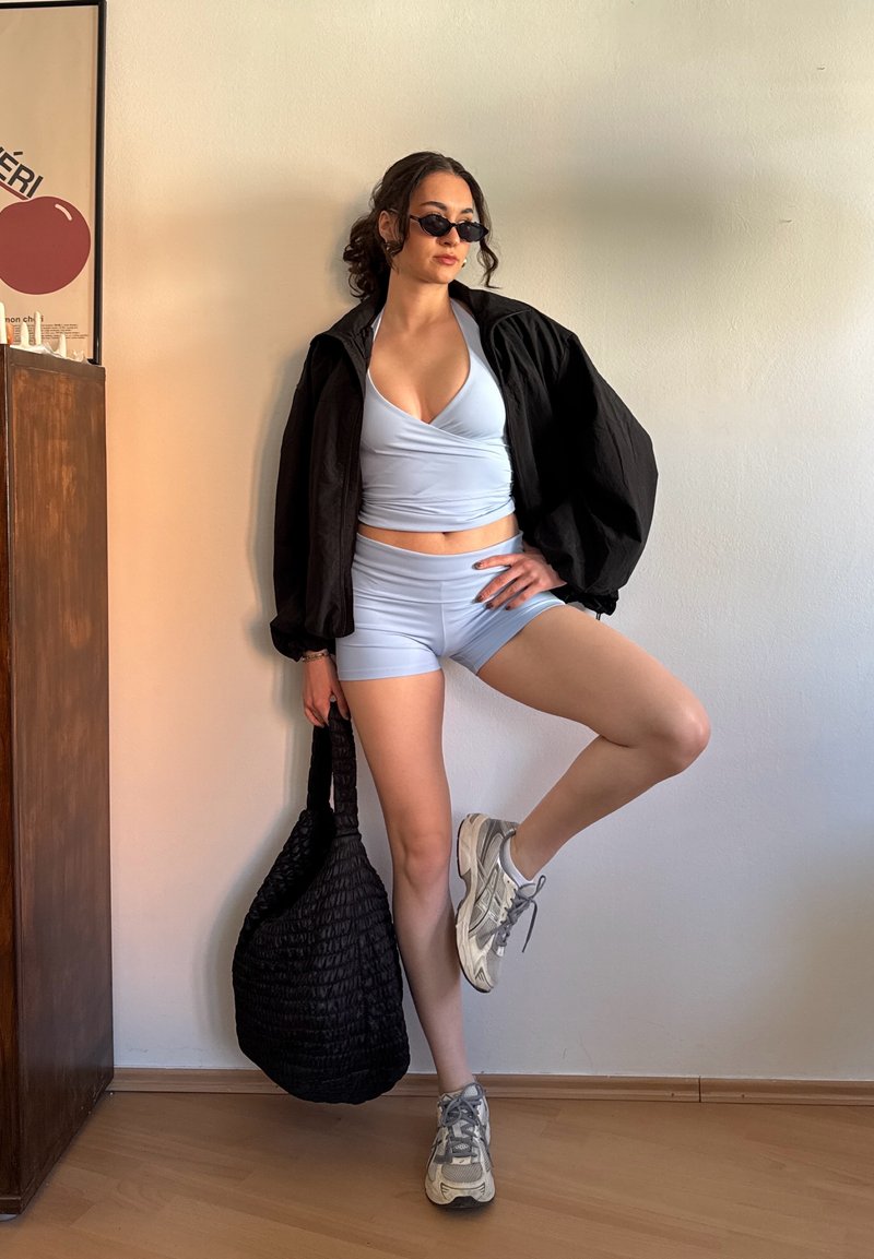 Femme portant un short de sport bleu clair et un haut avec une veste noire, des lunettes de soleil, tenant un sac noir, debout sur une jambe à l'intérieur près d'une armoire en bois.