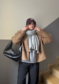Brauner Übergrößenmantel, grauer Schal, schwarze Tasche, grauer Pullover, schwarze Hose, kastanienfarbene Mütze mit Logo, gewellte Haare sichtbar, drinnen mit Treppe.
