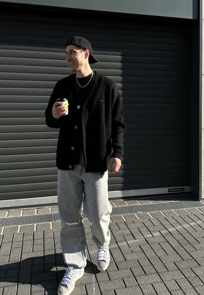 Cárdigan negro de canalé con escote en V, jeans gris de pierna ancha y zapatillas grises. Sosteniendo una taza de café. El fondo presenta una puerta de garaje gris.