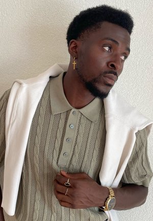 Light olive green knit polo shirt with textured pattern, short sleeves; worn with a white rolled sweater draped over shoulders, gold watch and ring.