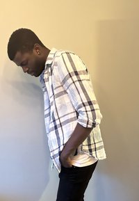 Chemise à carreaux claire en blanc avec des carrés bleus et noirs, portée par-dessus une chemise blanche, associée à un jean noir ; la personne est de profil contre un mur.