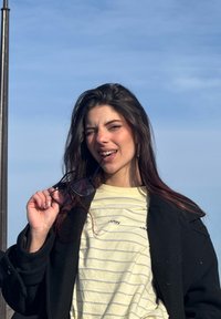 Jeune femme debout en plein air sous la lumière du soleil, faisant un clin d'œil et tenant des lunettes à monture noire près de la bouche, portant une chemise rayée jaune et un manteau noir.