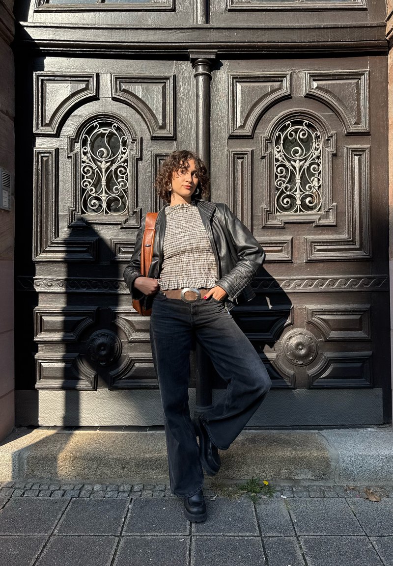 Casaco de cabedal preto sobre uma blusa xadrez, combinado com jeans pretos e botas robustas. O fundo apresenta portas de madeira ornadas.