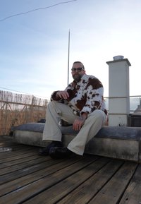 Homme portant des lunettes de soleil, un manteau en fourrure tacheté marron et blanc, un pantalon crème et des chaussures noires, assis sur une terrasse en bois sur un toit au coucher du soleil.