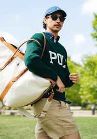 Mies, joka wear vihreä Polo-neule, sininen hattu, aurinkolasit ja vaaleanbeiget shortsit, kantaa suurta valkoista ja ruskeaa nahkatyylikassia ulkona.