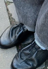 Chaussures en cuir noir texturé avec bouts arrondis, comportant une couture centrale et des lacets noués, associées à un pantalon gris et un tissu bleu clair apercevant.