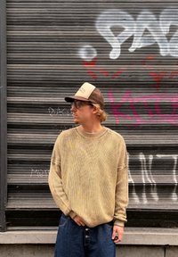 Smėlio spalvos megztinis megztas laisvo silueto su apsi�ūtais rankogaliais, derinamas su mėlynais plačiomis kelnėmis. Fone matyti pilkas žaliuzes ir grafitis.