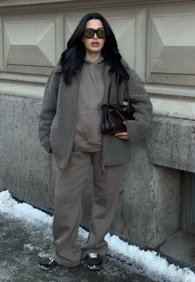 Mujer con cabello largo y negro que lleva una chaqueta de forro polar gris sobredimensionada, sudadera con capucha taupe y pantalones de chándal, zapatillas negras, gafas de sol, sosteniendo un bolso oscuro junto a una pared de piedra.